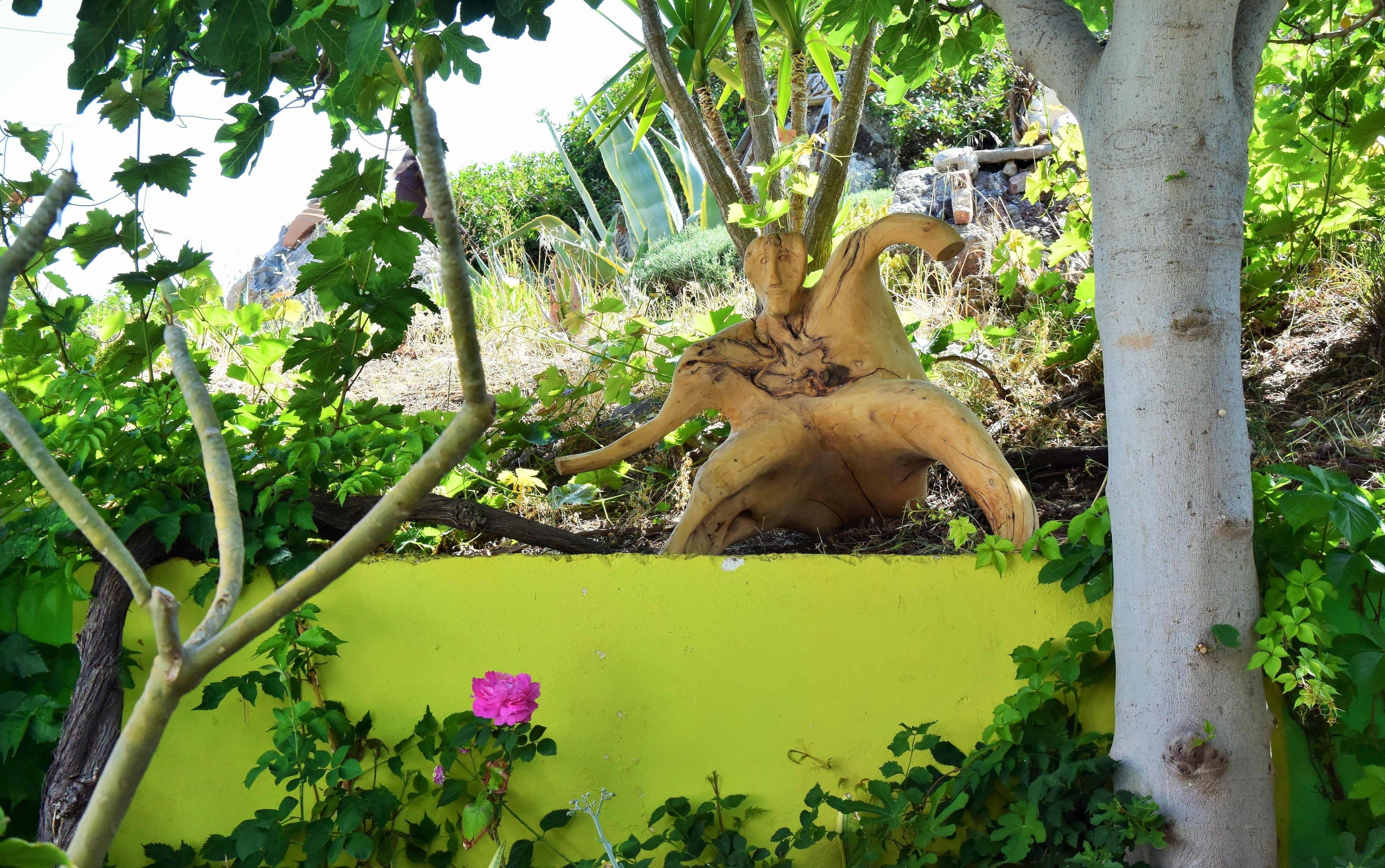 Un jardín con un tronco de árbol convertido en escultura, follaje verde y una pared amarilla.