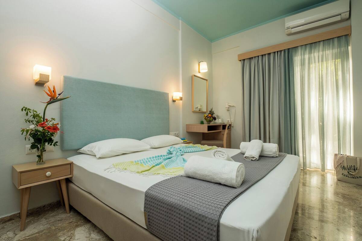 A hotel room with a bed, bedside table, nightstand, mirror, and a window with curtains.