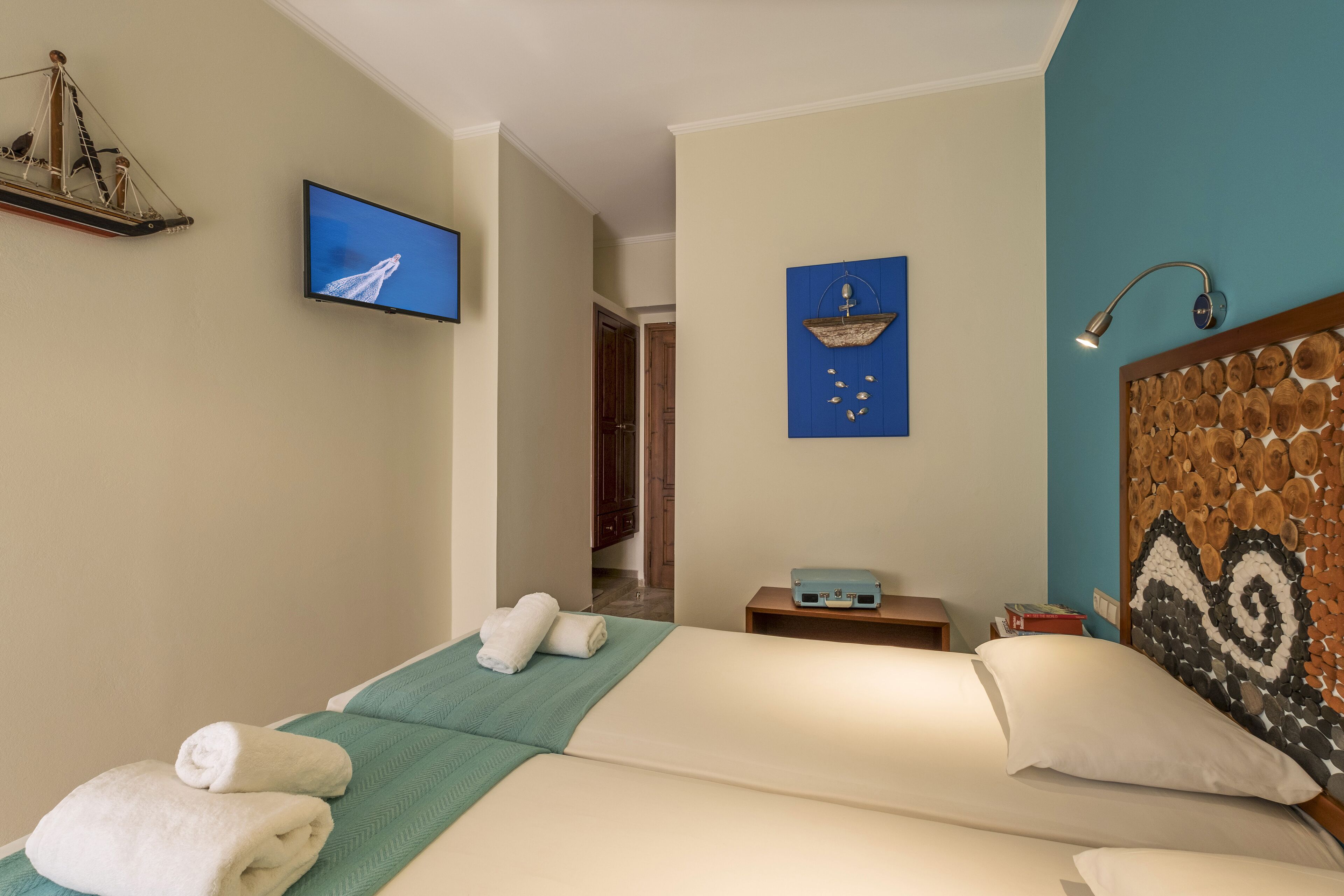 A hotel room with a bed, a television, a wall-mounted shelf with a ship model, a wall art piece, and a wooden headboard.