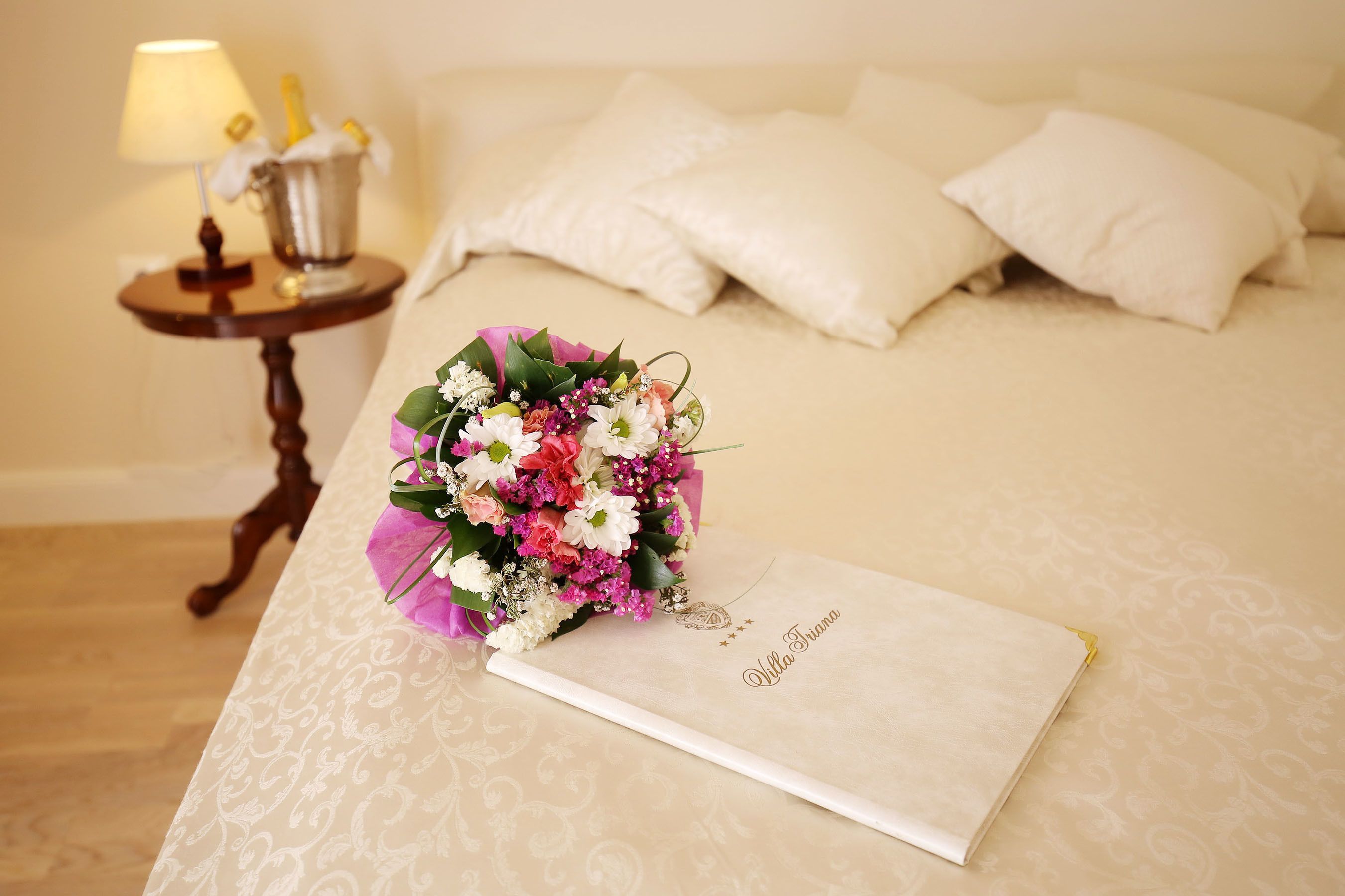 Una habitación ordenada con una cama, una mesita de noche con una lámpara y un ramo de flores con una tarjeta.