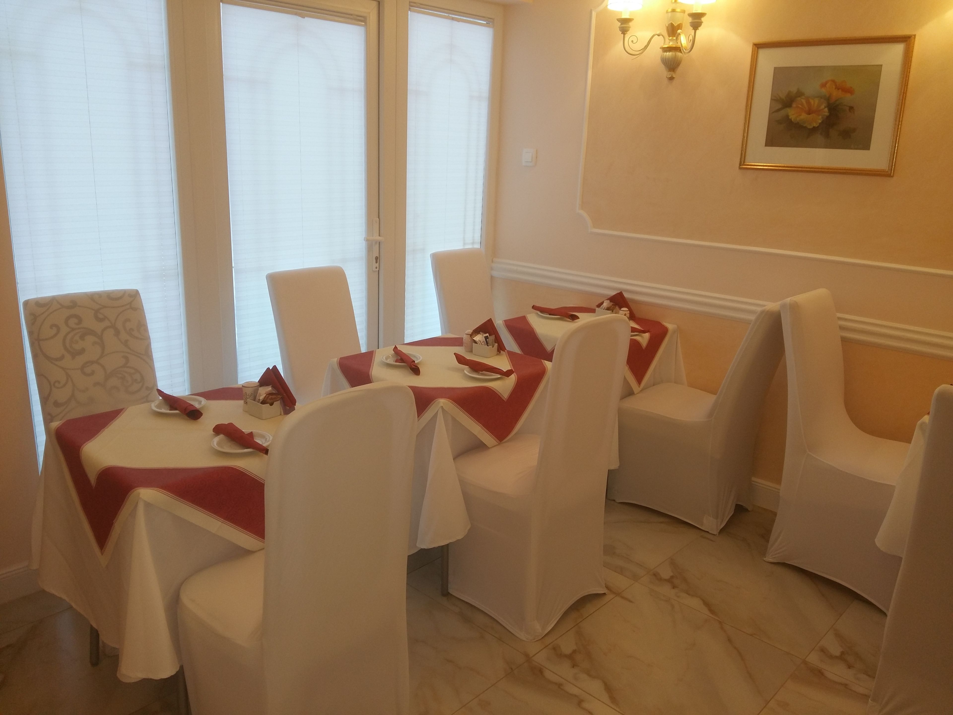 A dining area with white chairs and tables, each with a red tablecloth. There are two wall-mounted light fixtures and a framed picture on the wall.