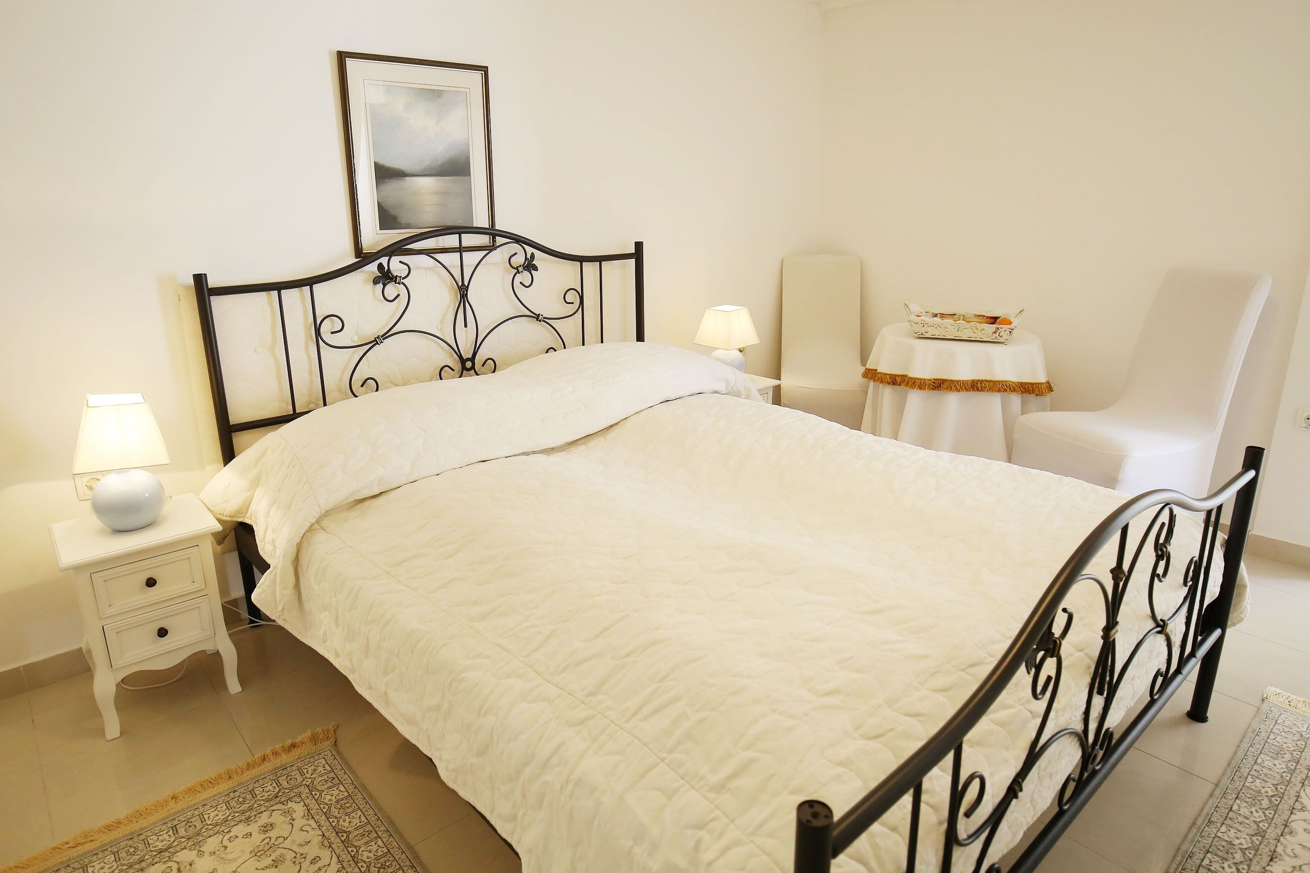 A neatly made bed with a white quilt, a bedside table with a lamp, a chair, and a small table with a decorative item.