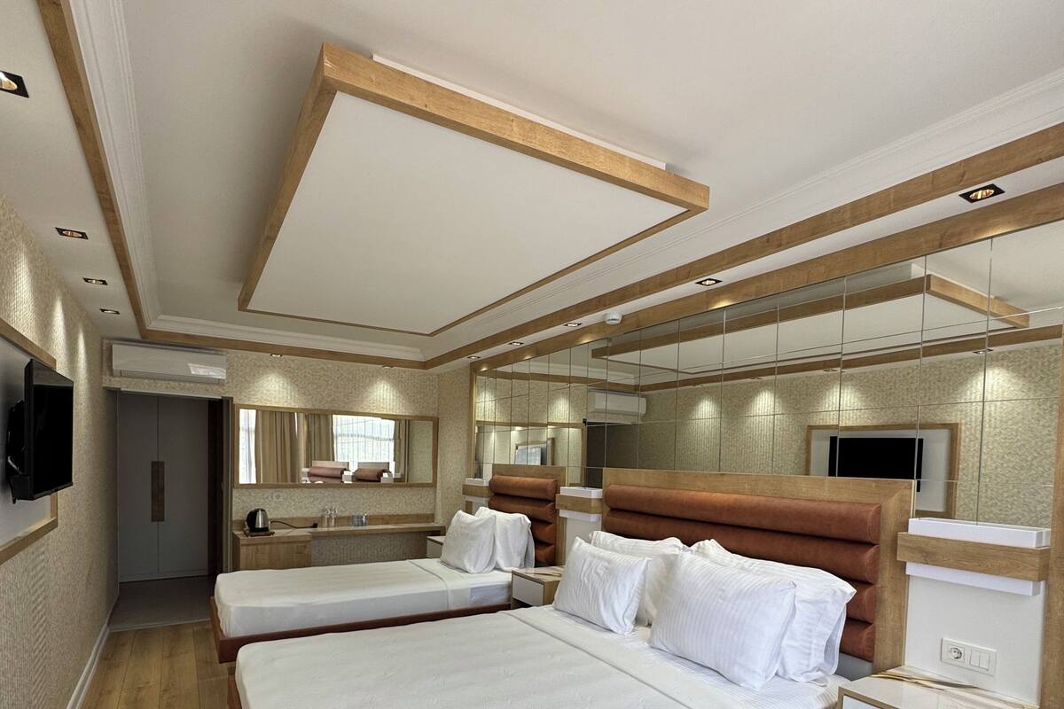 A hotel room with a large bed, a bedside table, a round wooden table with fruit and a bottle, and a TV mounted on the wall.