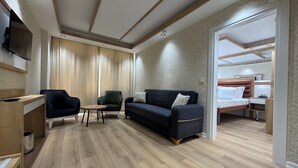 A modern hotel room with a wooden floor, a dark blue sofa, a green armchair, a small round table, and a bed with a wooden headboard. - Alican Hotel (Izmir)