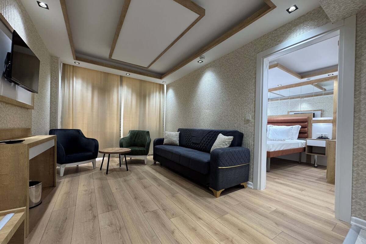 A modern hotel room with a wooden floor, a dark blue sofa, a green armchair, a small round table, and a bed with a wooden headboard.