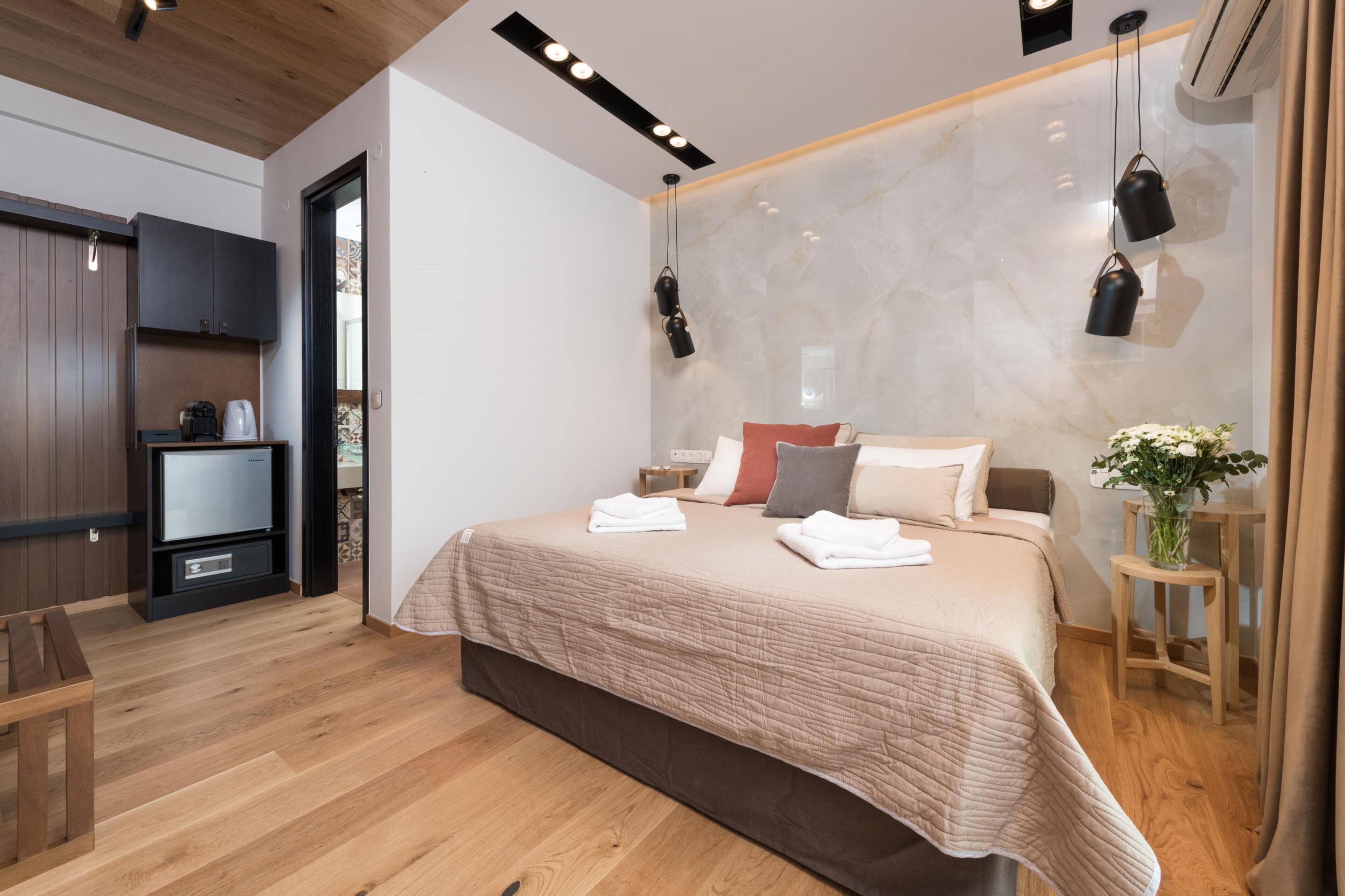 A modern bedroom with a wooden floor, a bed with a beige quilt, a nightstand, a small table with a vase, and a wardrobe.