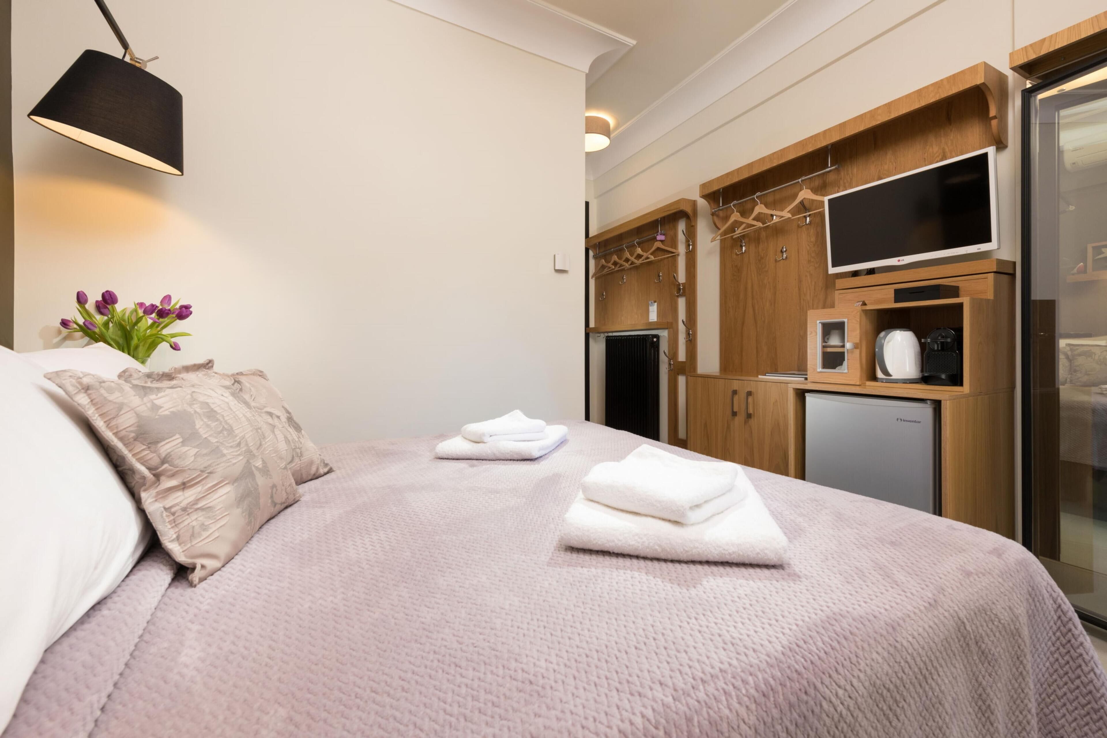 A neatly made bed with a pink blanket, a wooden cabinet with a TV, and a small kitchenette.