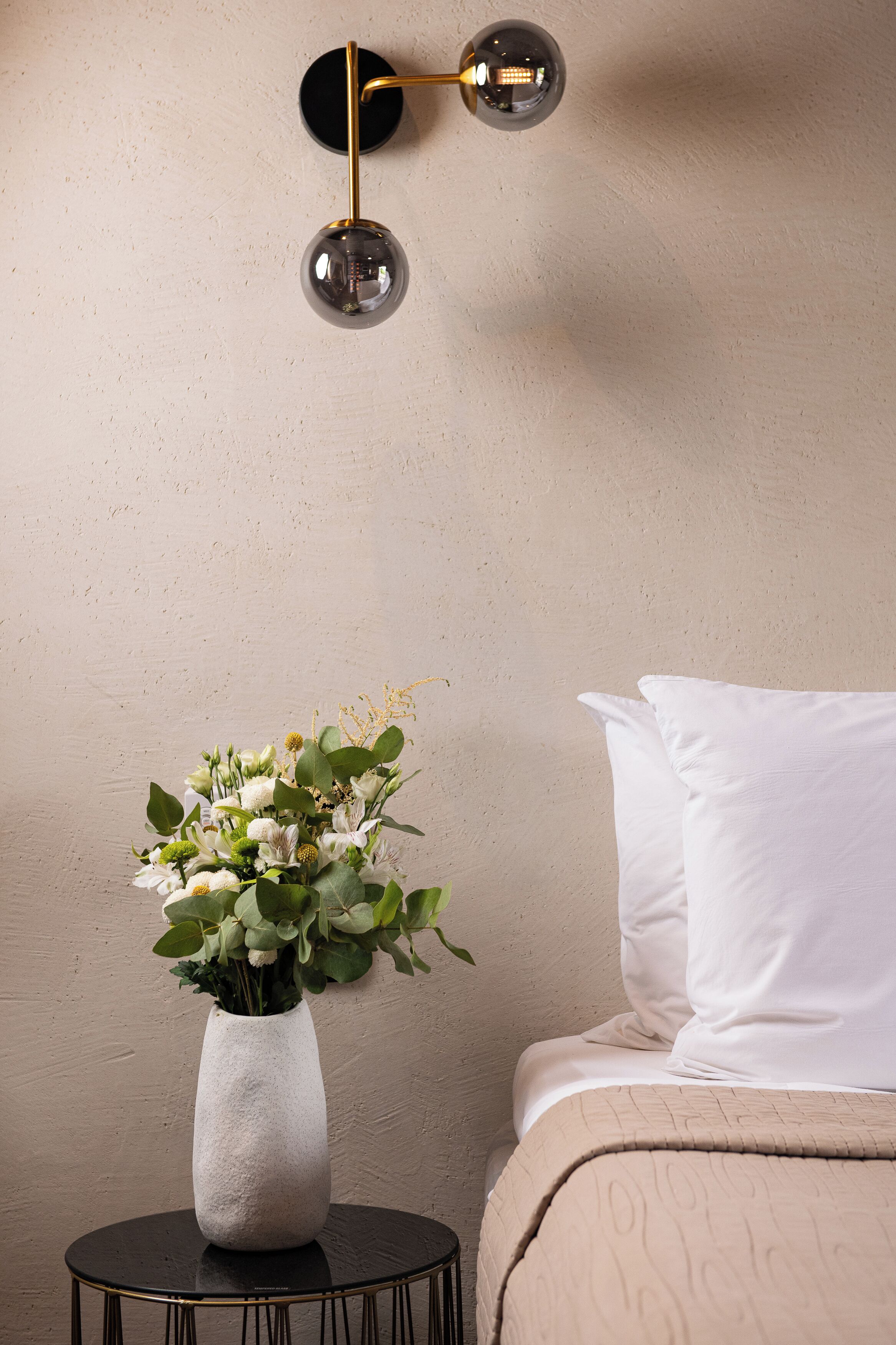 Una camera da letto con un letto, un comodino con un vaso di fiori e una lampada a parete.