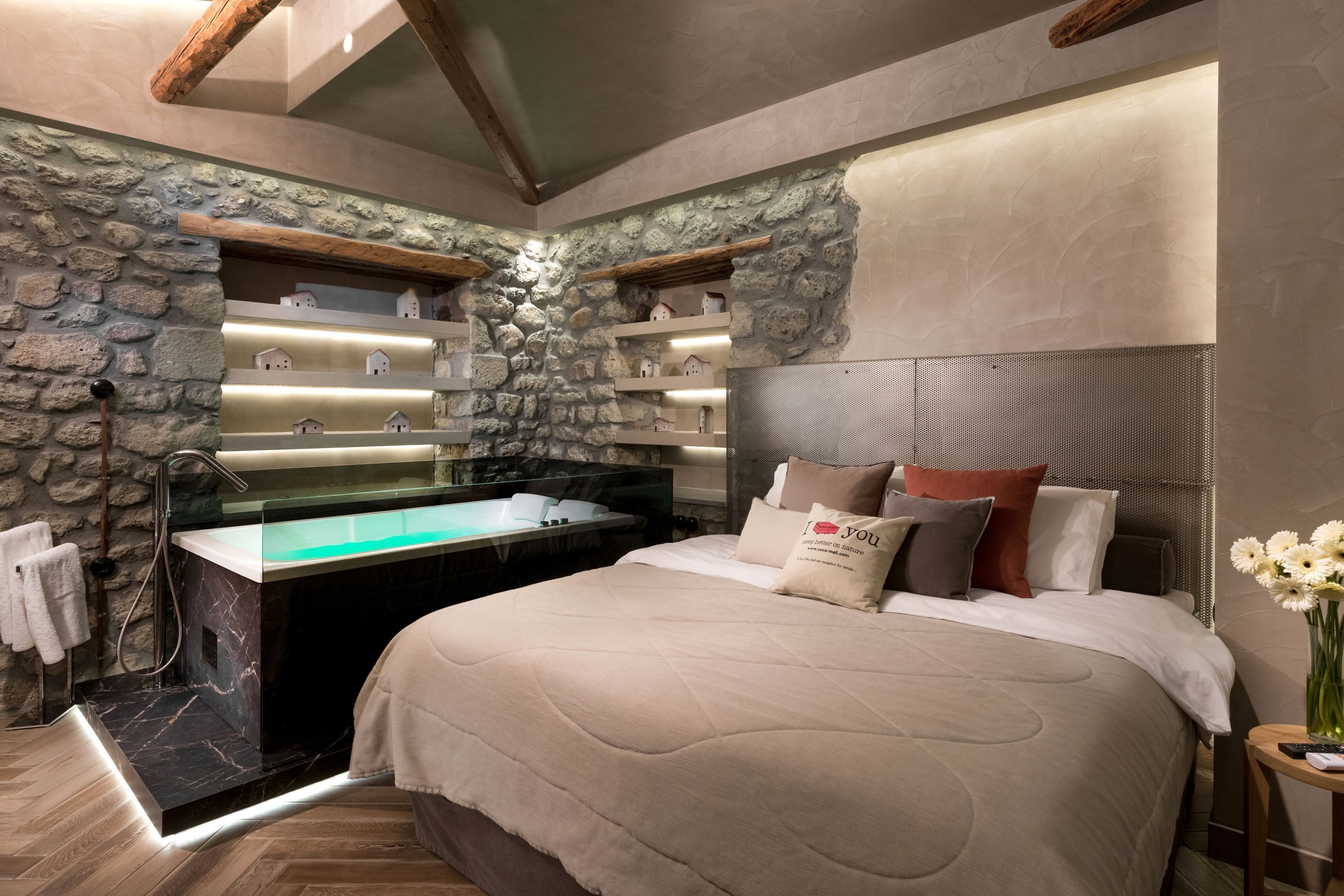 Une chambre moderne avec un mur d’accent en pierre, un grand lit, une baignoire intégrée et une petite table sur laquelle est posé un vase de fleurs.