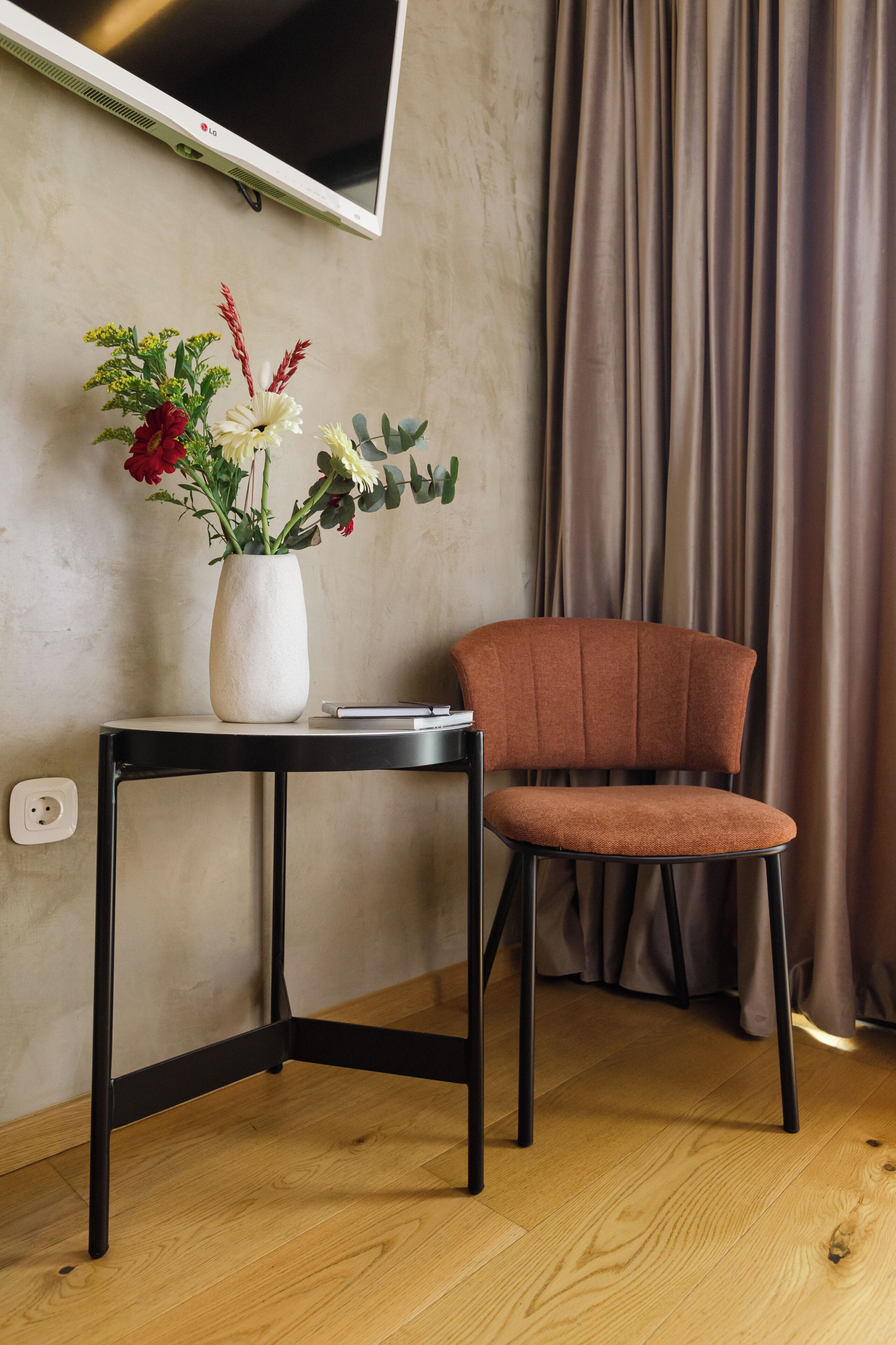 A room with a wooden floor, a black metal table with a white vase containing flowers, a brown upholstered chair, a black TV mounted on the wall, and a curtain.