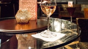 A glass of white wine on a round table with a menu and a decorative candle in a warmly lit room with large windows and classic decor. - Gallery Park Hotel & SPA, a Châteaux & Hôtels Collection (Riga)