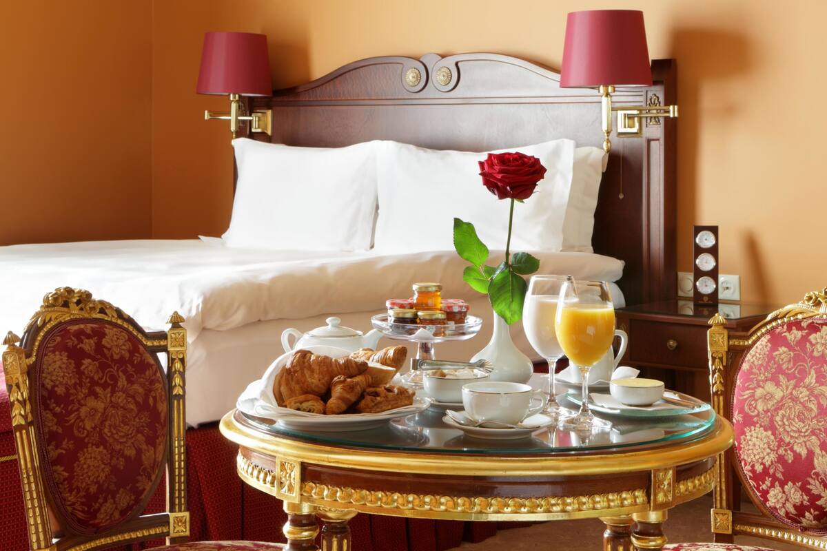 A hotel room with a neatly made bed, a breakfast table with croissants, a vase with a rose, and a bedside table with a clock and a lamp.