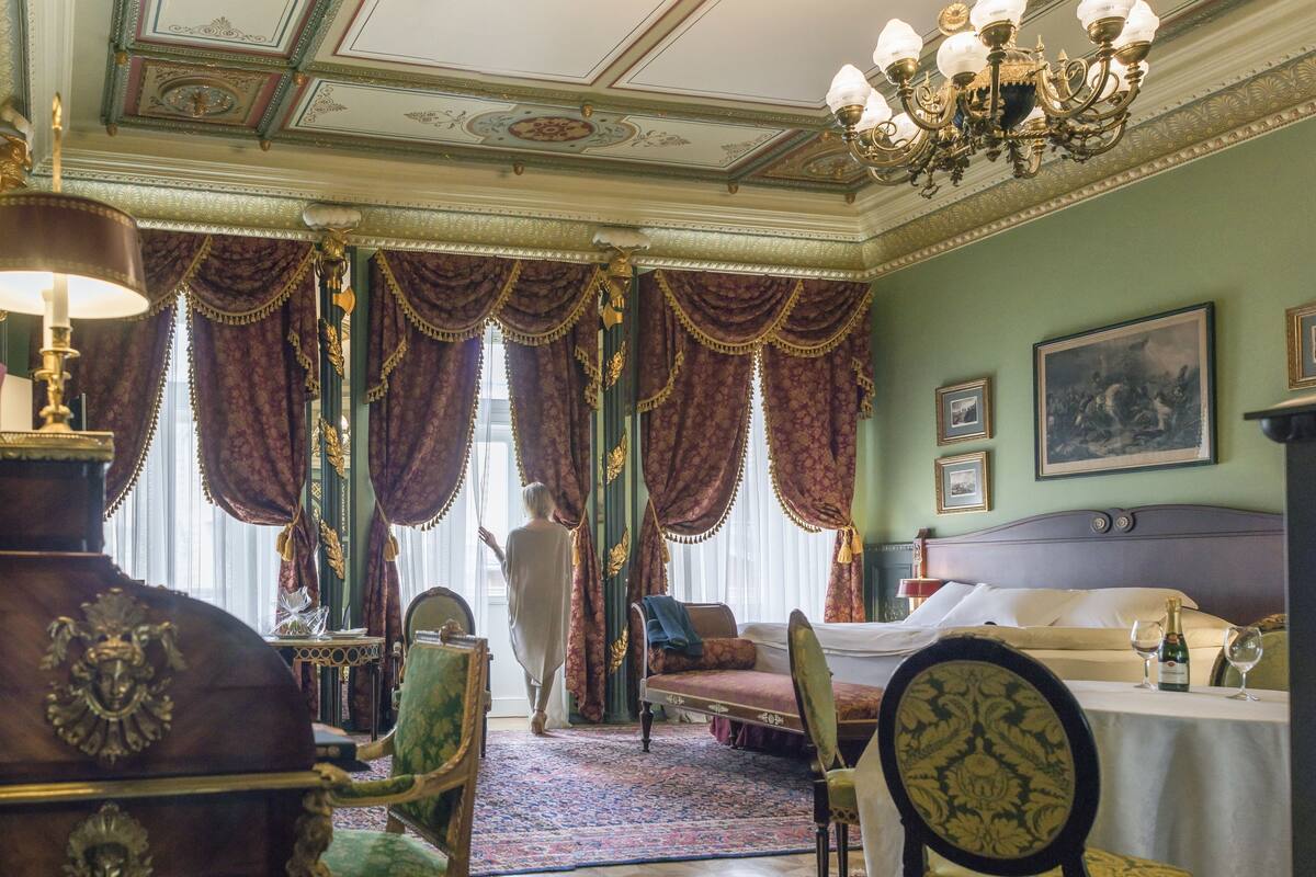 A room with a bed, a table with glasses, a chair, a chandelier, and a large window with curtains.