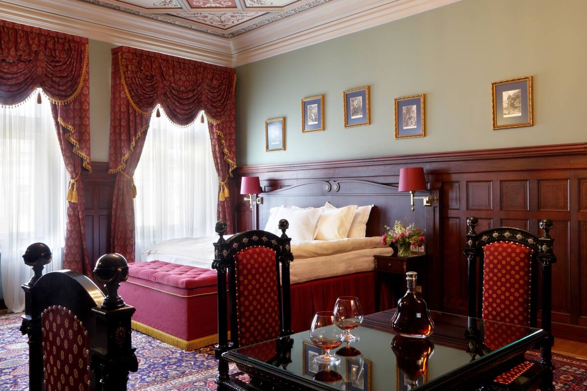A bedroom with a large bed, two chairs, a table with glasses, and framed pictures on the wall.