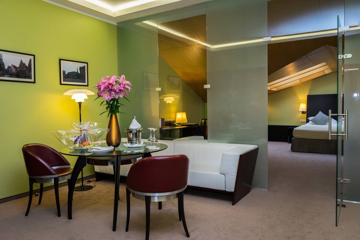 A hotel room with a glass partition, a dining area with a table and chairs, a white sofa, a bed, and a staircase leading to an upper level.