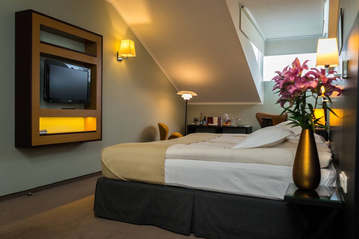 A hotel room with a bed, a television mounted on the wall, a desk with a lamp, and a vase with flowers.