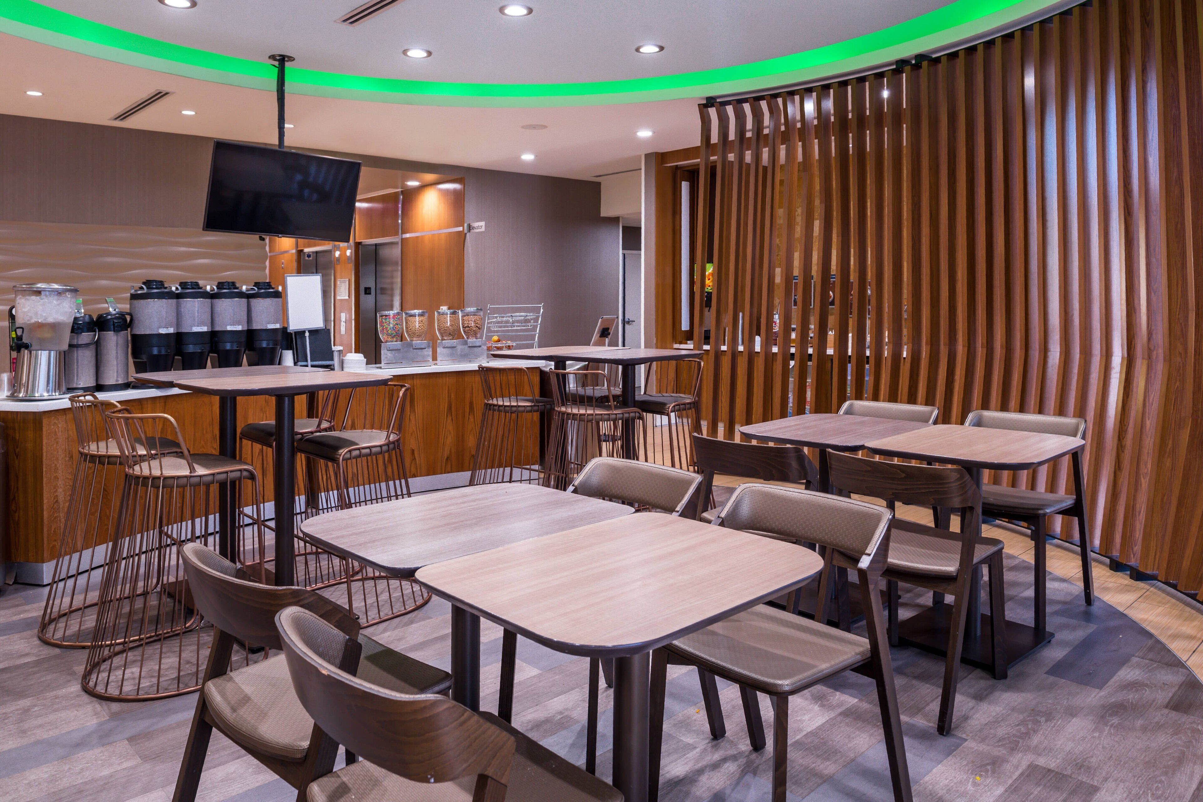 A modern restaurant with multiple tables and chairs, a counter with a coffee machine, and a television mounted on the wall.