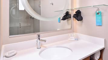 Un baño moderno con un espejo grande, un lavabo blanco y un estante de vidrio que sostiene una bolsa negra y una etiqueta azul.