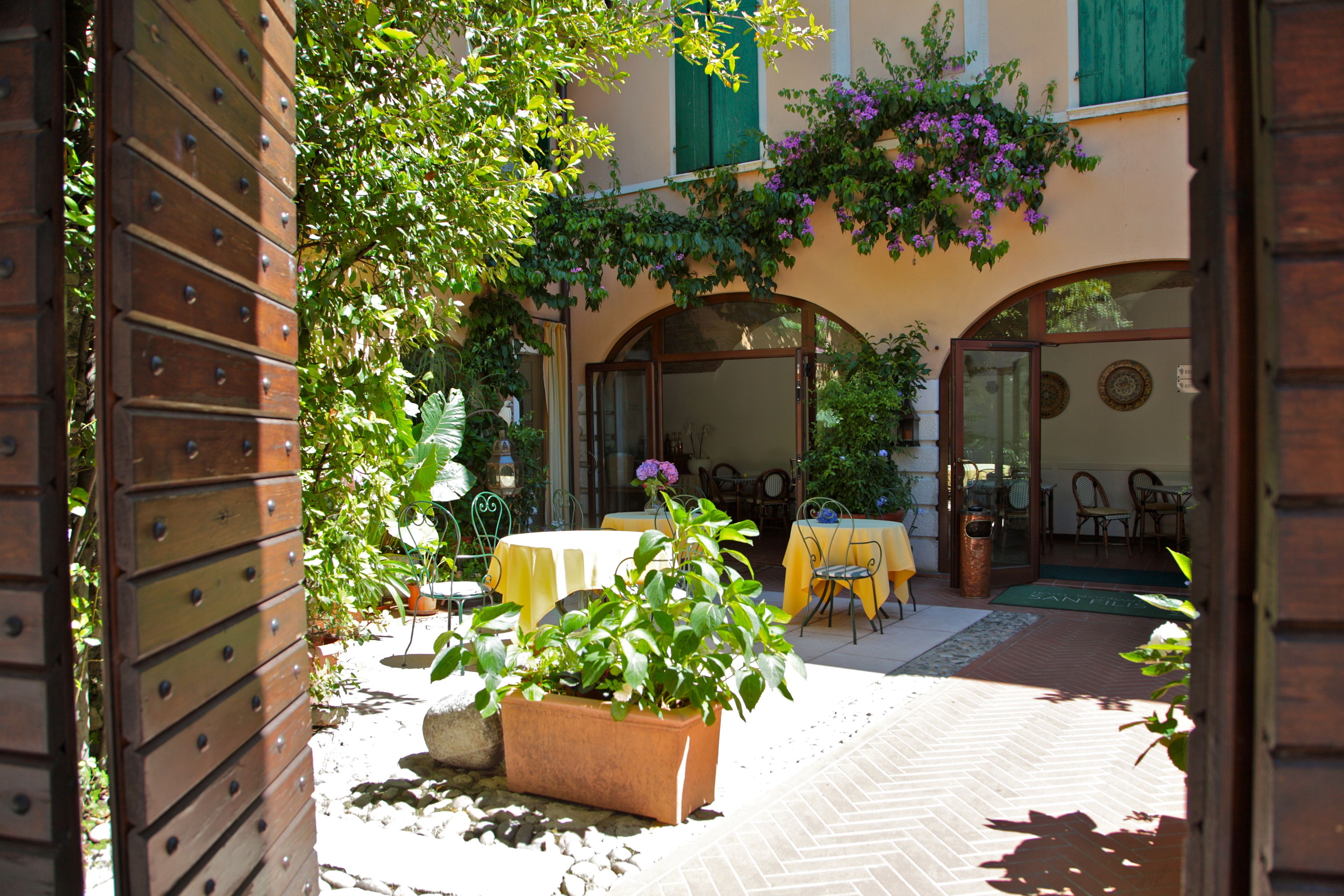 An outdoor patio with a brick floor, yellow tables, and green chairs. There are potted plants and a wooden gate.