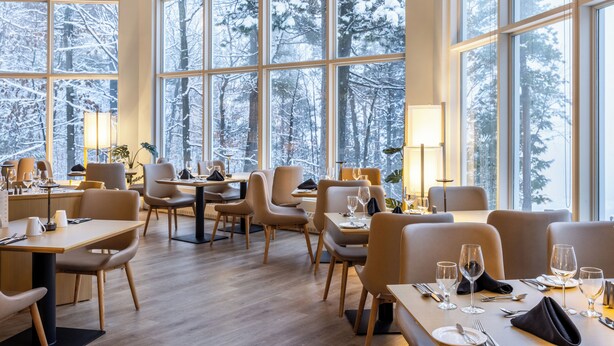 A restaurant with large windows offering a snowy forest view. Tables are set with wine glasses and cutlery.
