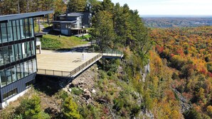 A modern house with a glass facade and a wooden deck overlooking a forested valley.