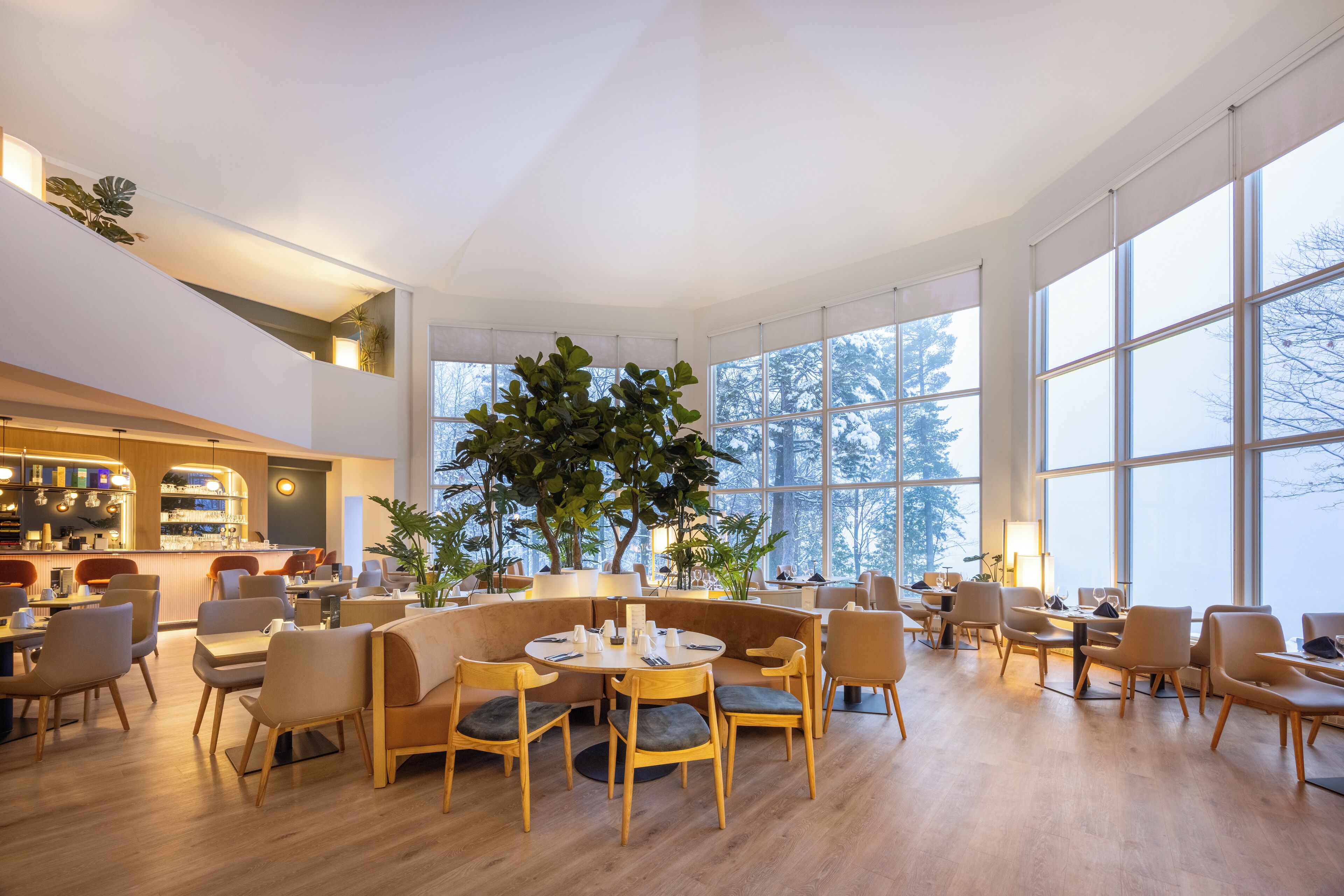 A spacious, well-lit indoor area with large windows, wooden flooring, and a variety of seating arrangements including round tables and chairs.