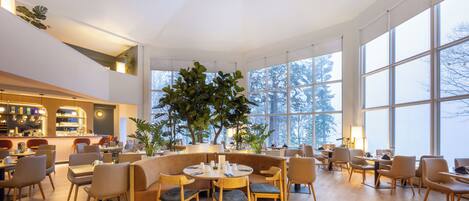 A spacious, well-lit indoor area with large windows, wooden flooring, and a variety of seating arrangements including round tables and chairs.