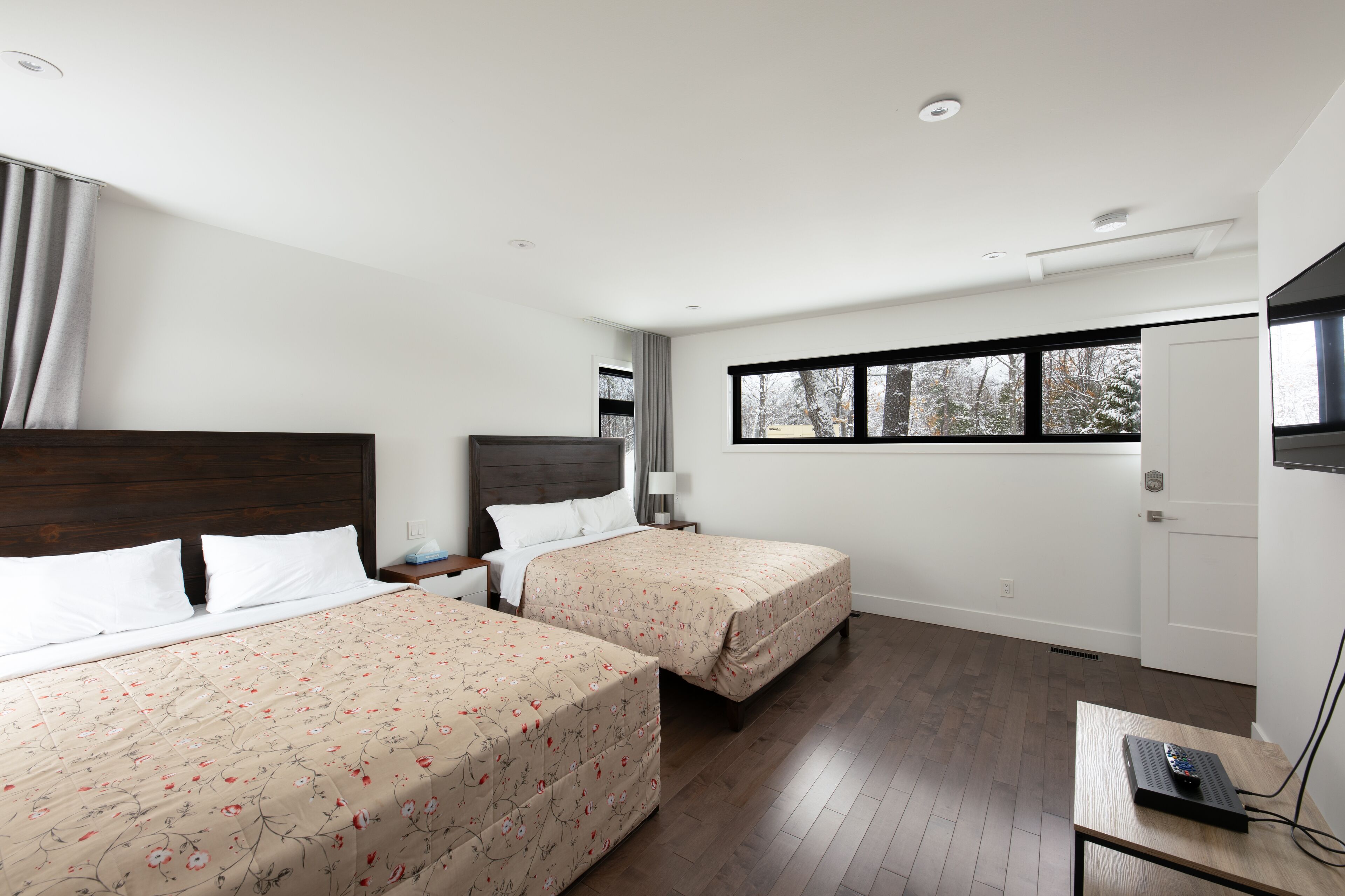 Quarto de hotel com duas camas, cabeceira de madeira, janela com vista para árvores e uma escrivaninha com controle remoto.