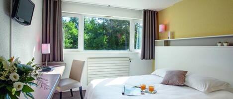 Habitación de hotel con cama, escritorio, silla, televisor, ventana con cortinas y mesita de noche con un tray con jugo de naranja.