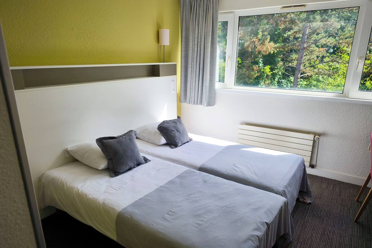 A hotel room with two beds, a window with a view of trees, a lamp, and a radiator.