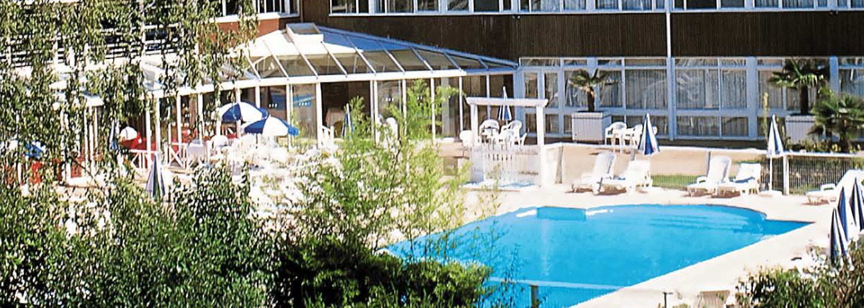 Piscina com espreguiçadeiras e guarda-sóis, rodeada por vegetação e um edifício com grandes janelas.