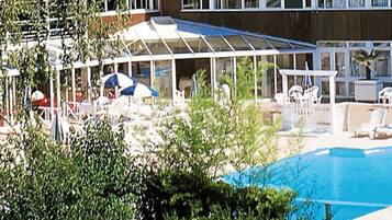 Piscina com espreguiçadeiras e guarda-sóis, rodeada por vegetação e um edifício com grandes janelas.