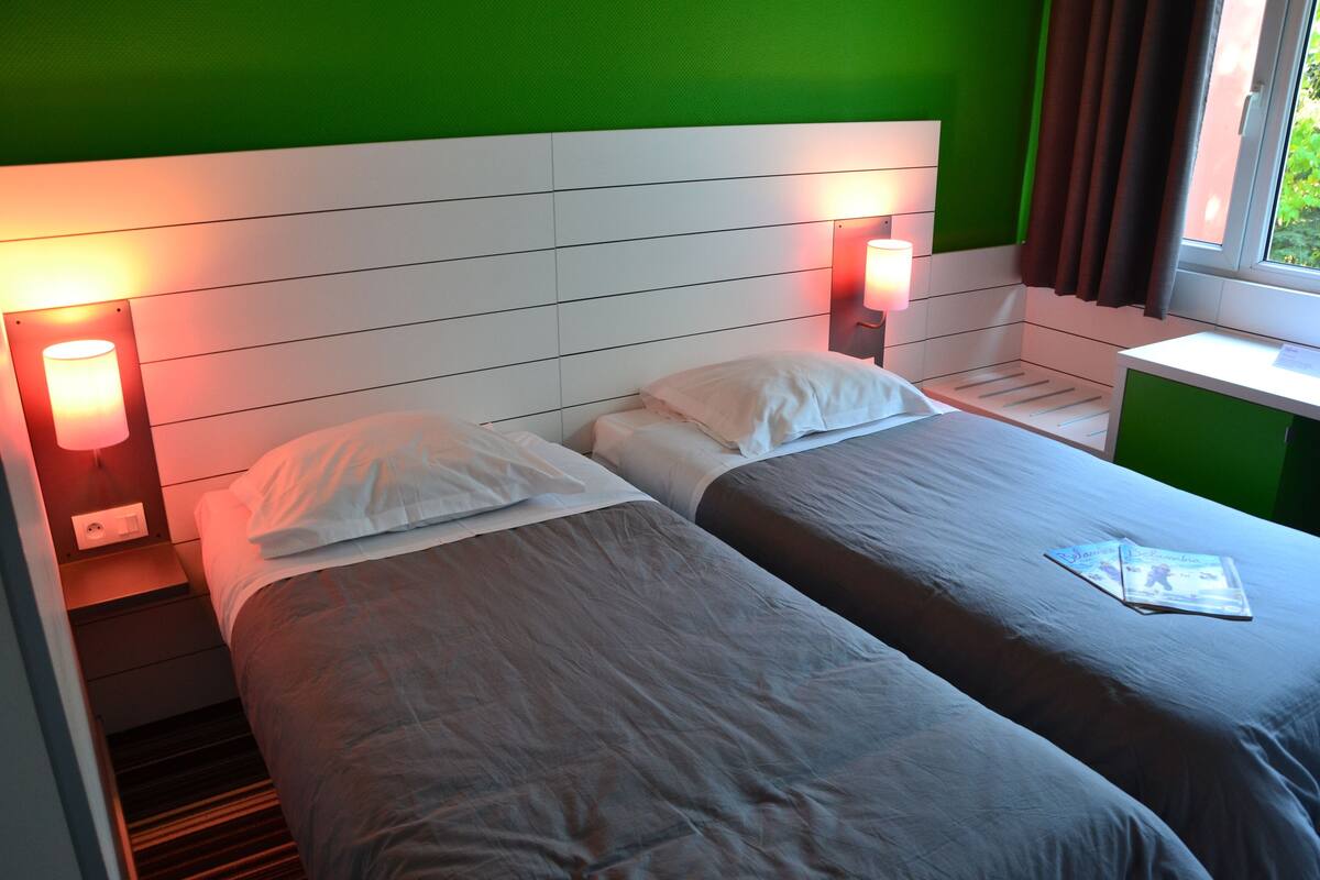 A double bed with white linens, two bedside lamps, a window with curtains, and a green wall.
