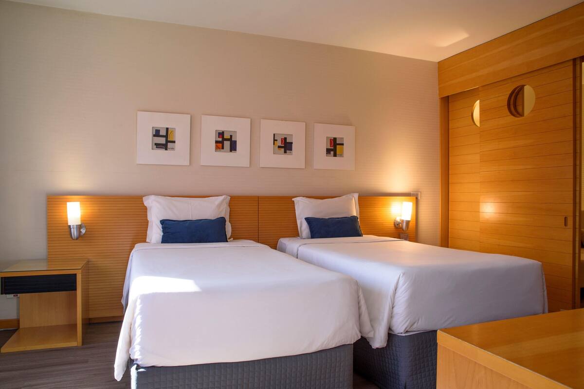 Habitación de hotel con dos camas, cabecero de madera, mesitas de noche con lámparas y cuadros en la pared.