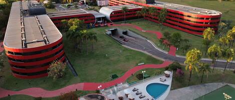 A modern hotel with multiple red and black buildings, a central courtyard with palm trees, and a swimming pool area.