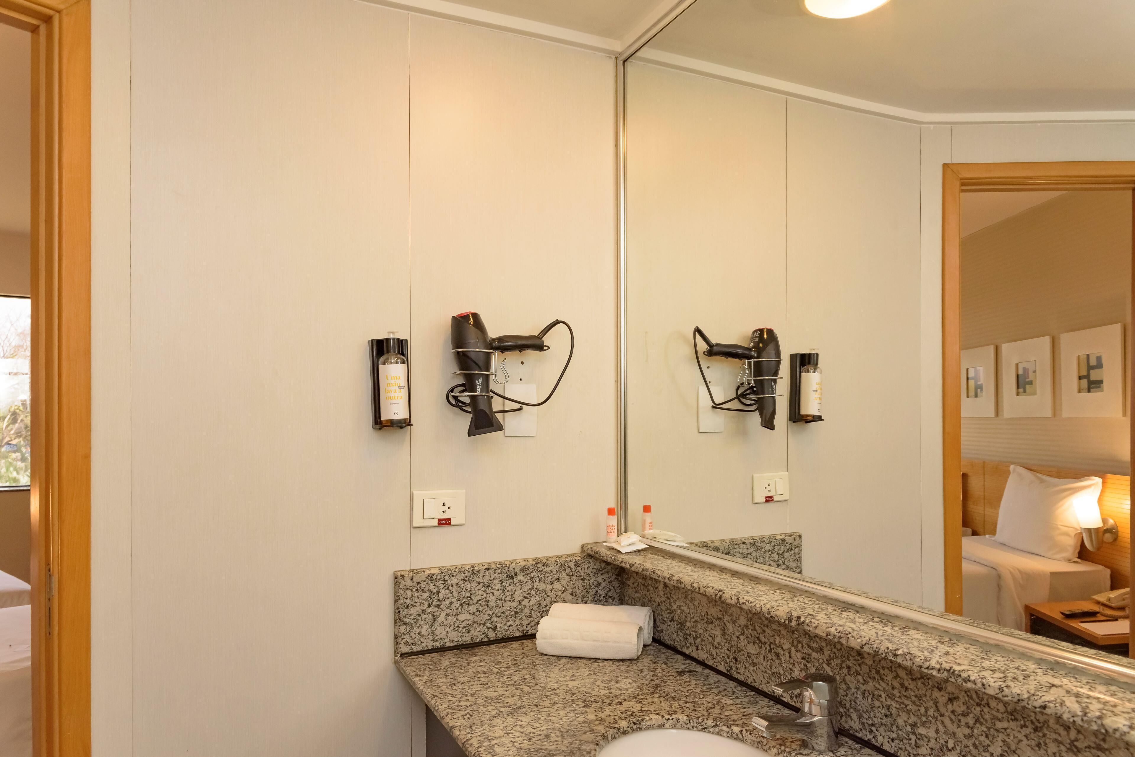 Baño con encimera de granito, lavabo, espejo y dos secadores de pelo fijados a la pared.