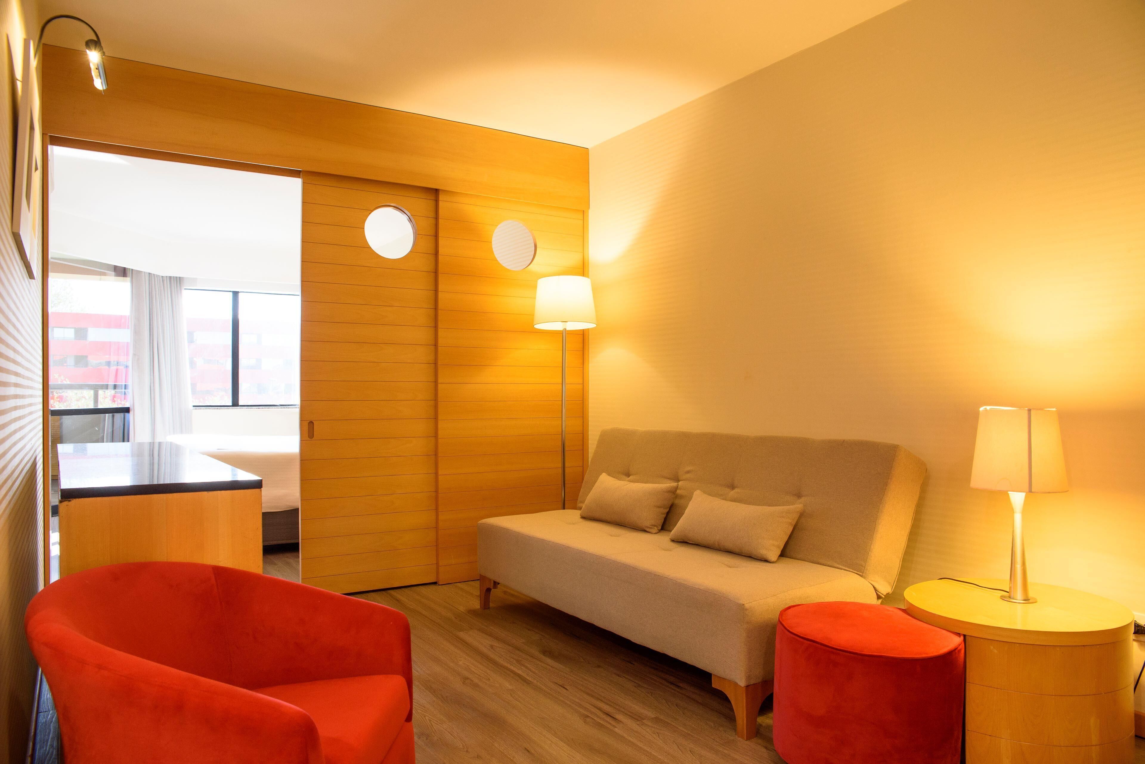 Une chambre d’hôtel moderne avec un canapé, deux fauteuils rouges, une petite table et une lampe.