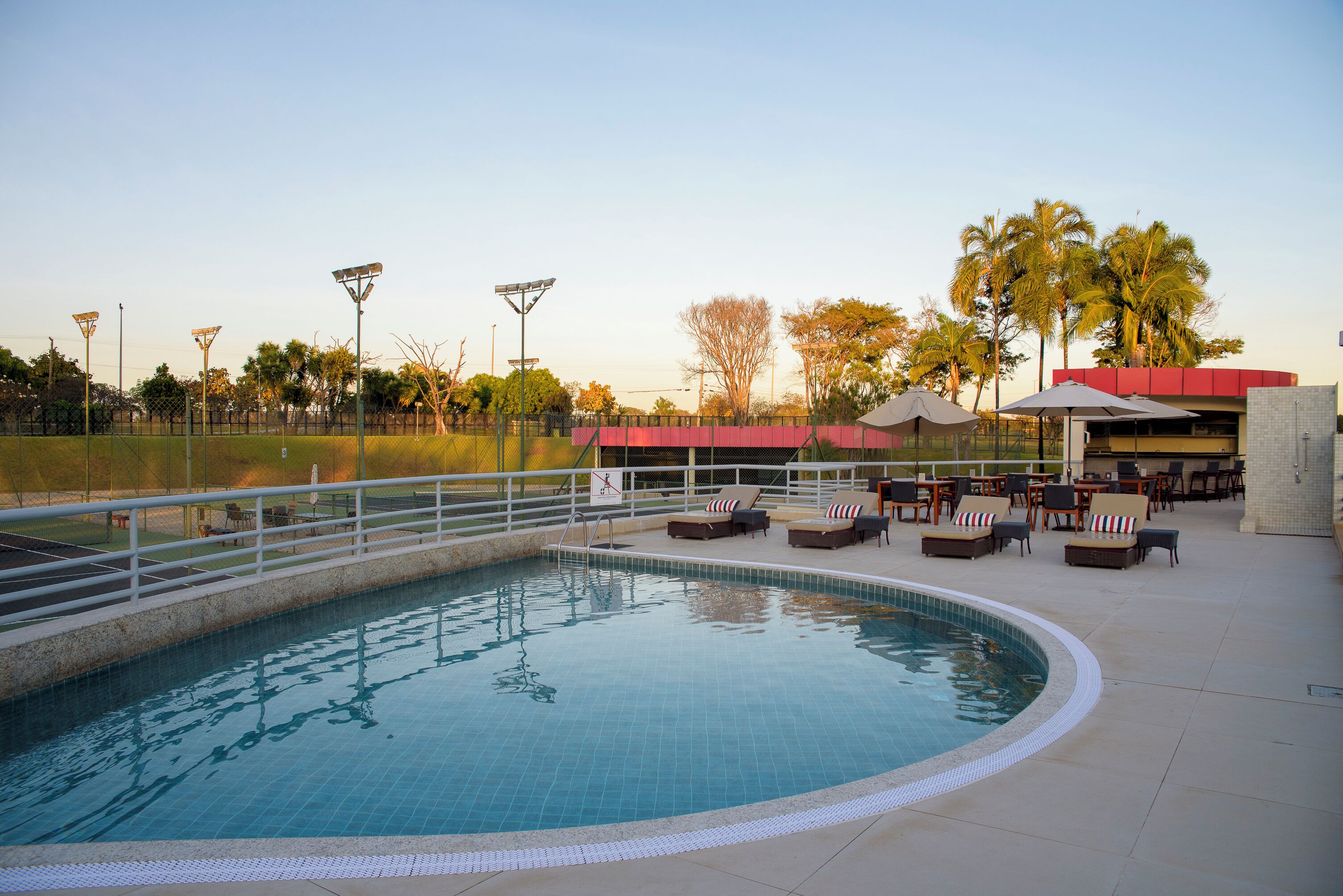 Piscina com cadeiras de sol e guarda-sóis, cercada por uma vedação e palmeiras.
