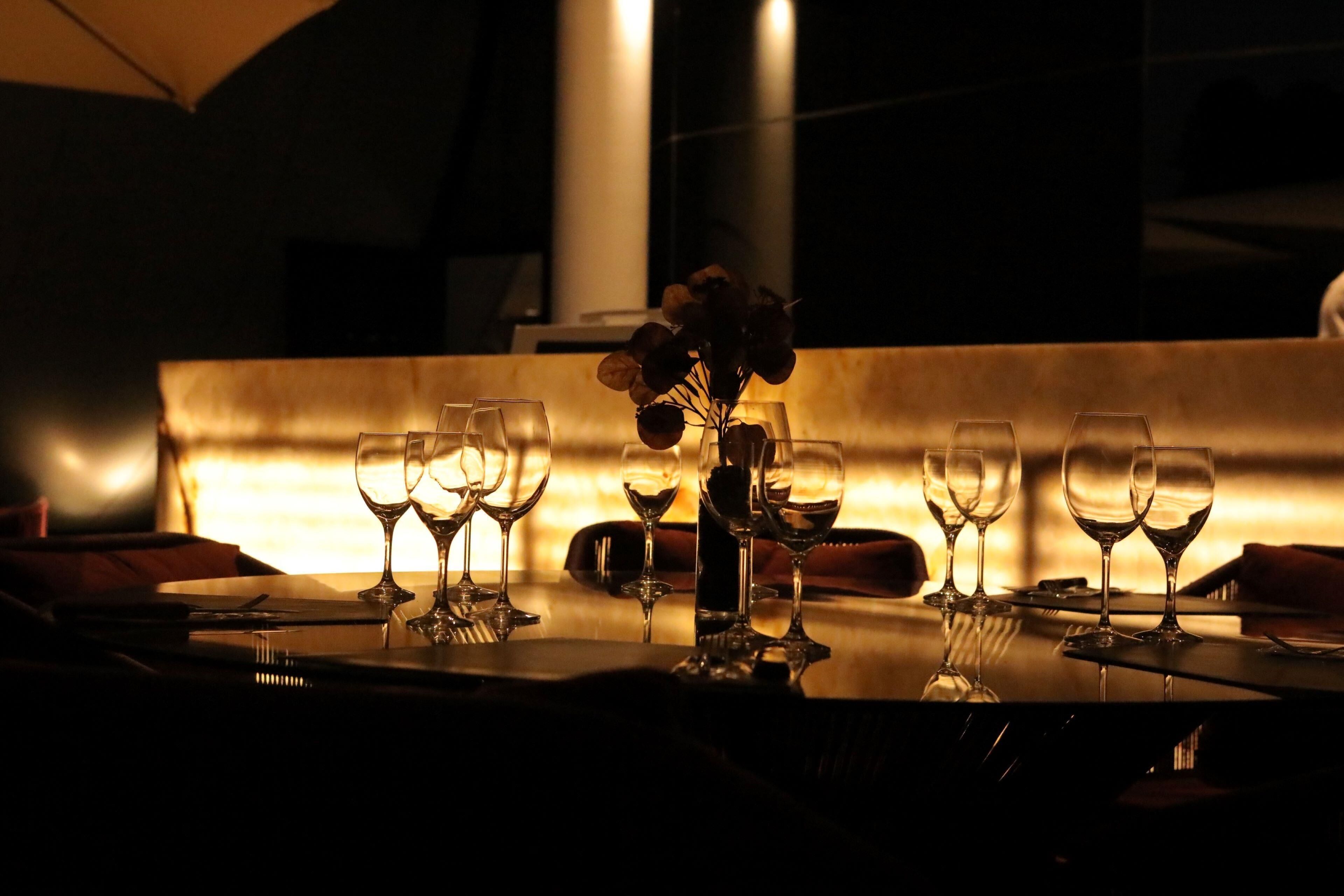 A dimly lit dining area with a table set for a meal, featuring a vase with dried flowers and several wine glasses.