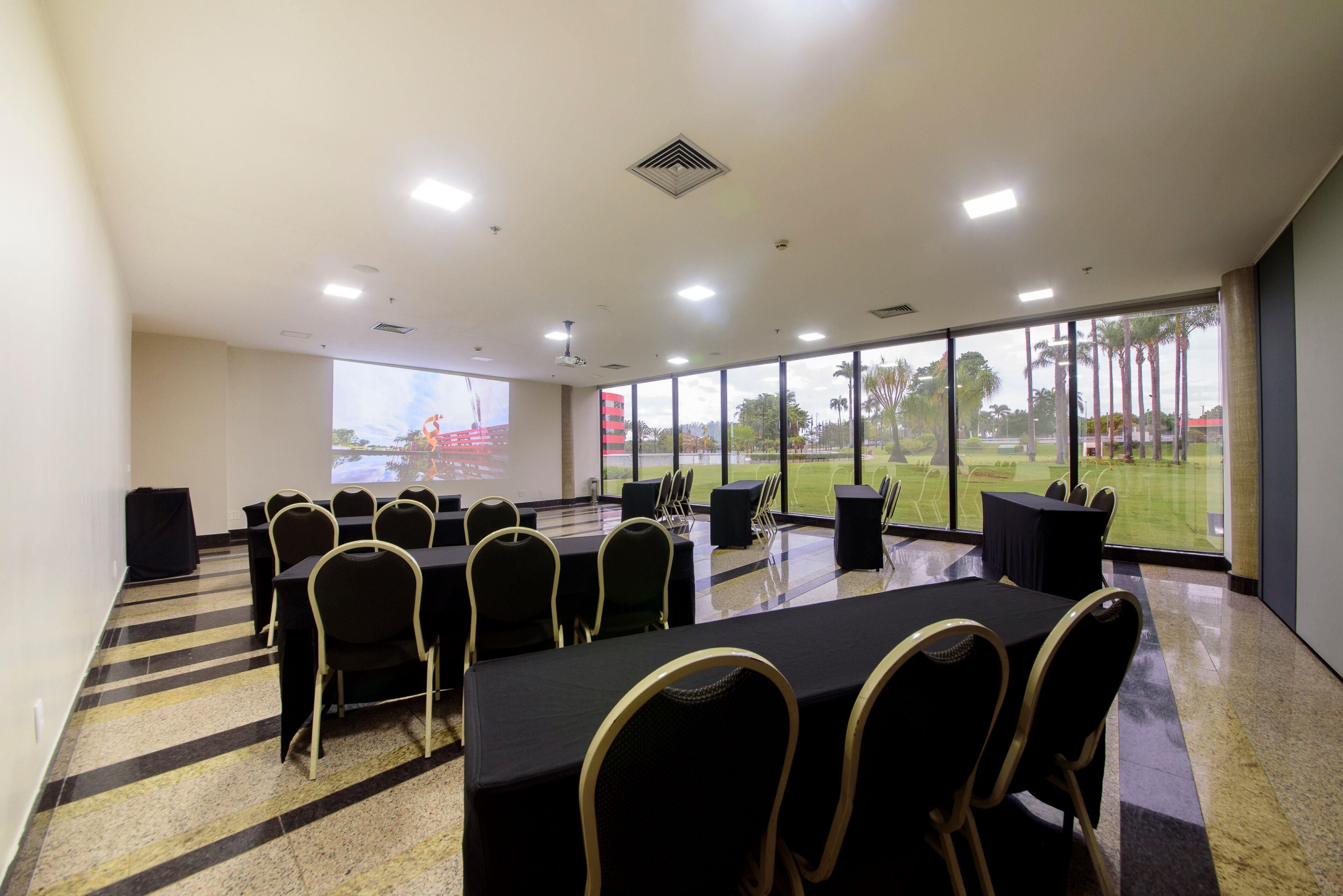 Sala de conferências com mesas e cadeiras, amplas janelas com vista para palmeiras e um campo de esportes, e uma tela de projetor.