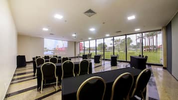 Sala de conferências com mesas e cadeiras, amplas janelas com vista para palmeiras e um campo de esportes, e uma tela de projetor.