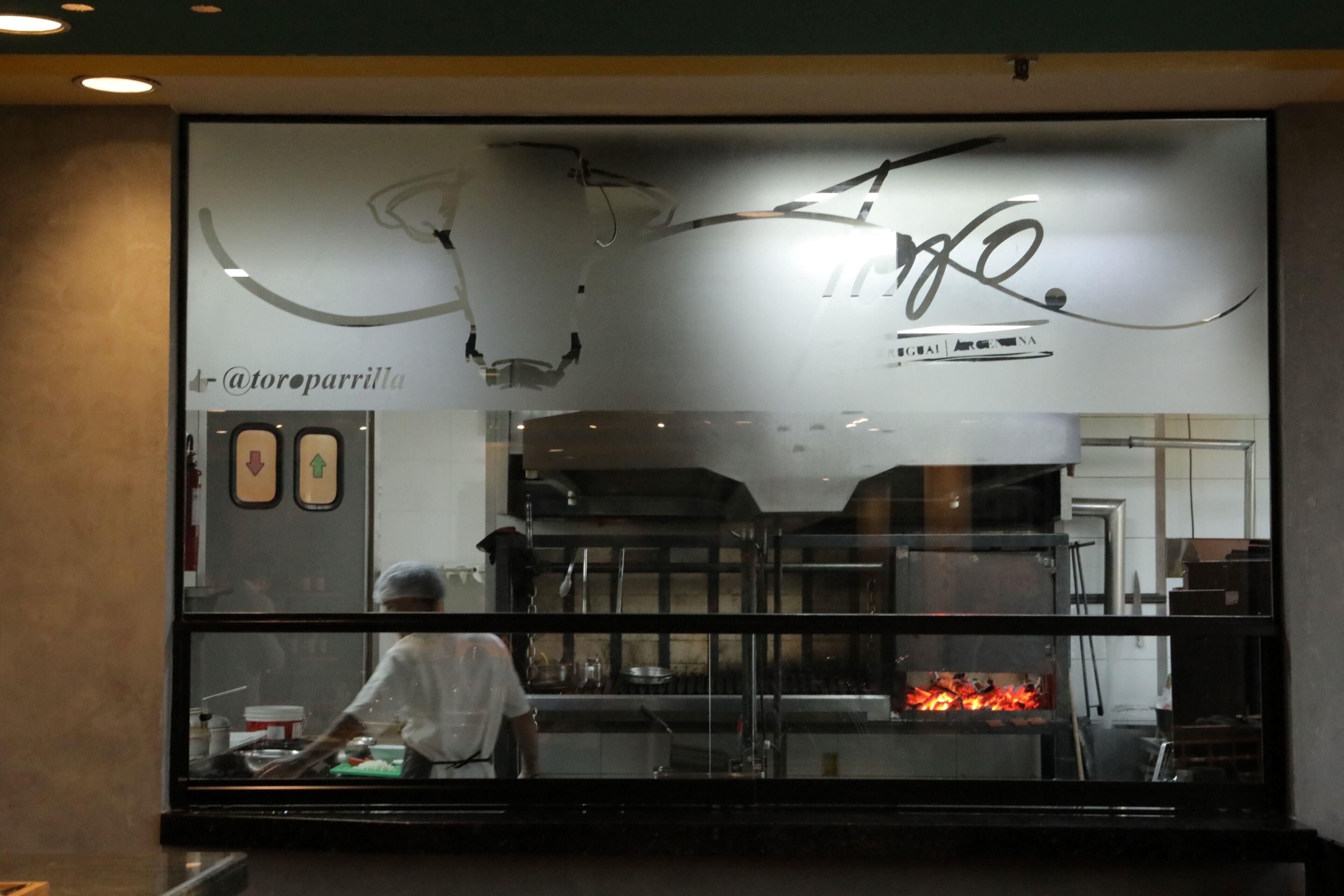 Un restaurant doté d’une grande baie vitrée, où l’on peut voir un chef préparer des plats et un four dont on aperçoit les flammes.