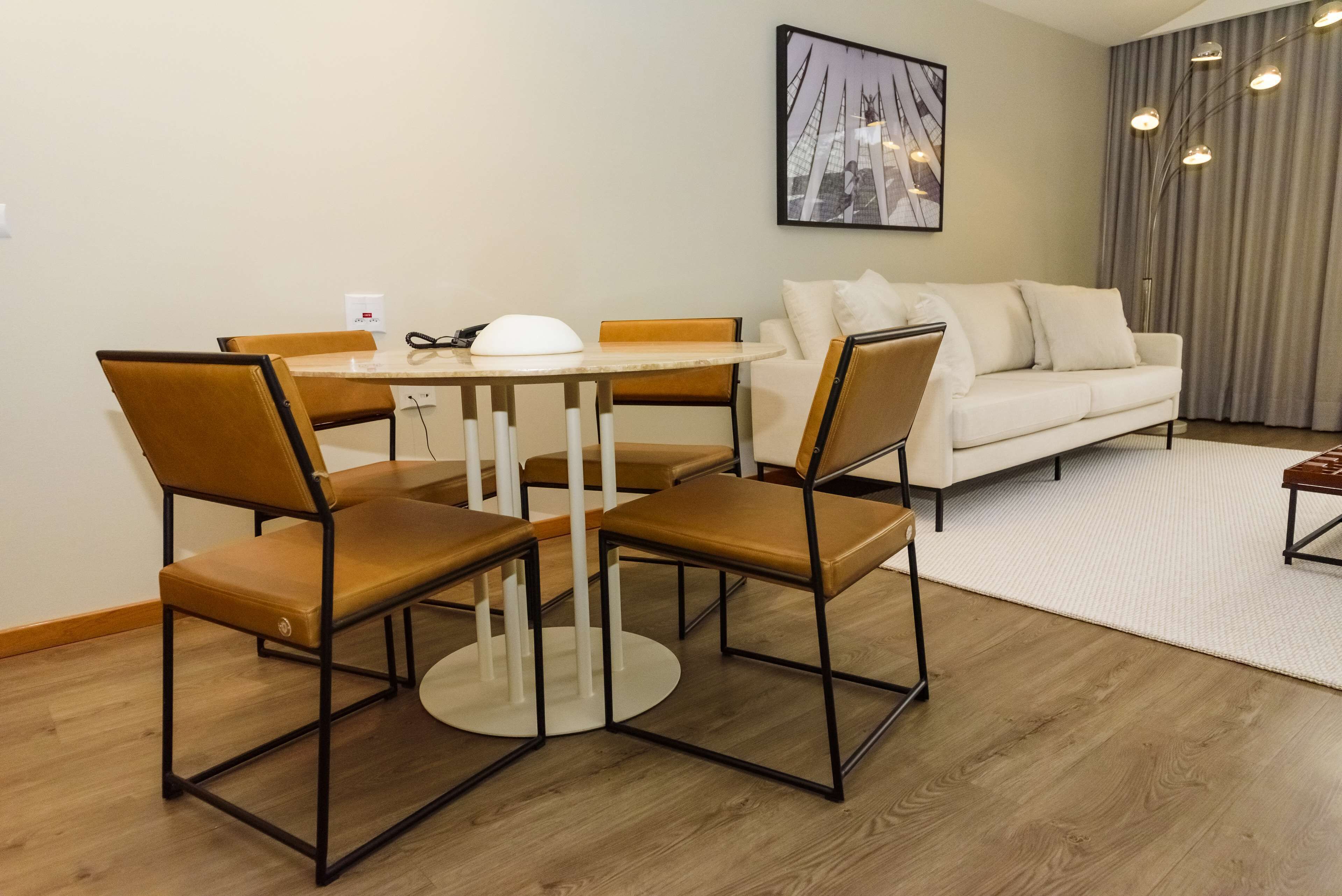 A modern living room with a round dining table, four brown chairs, a white sofa, and a large window with curtains.