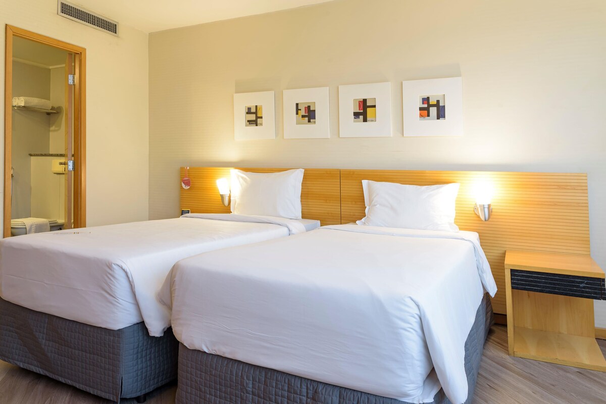Une chambre d’hôtel avec deux lits, une tête de lit en bois, des œuvres d’art accrochées au mur et une table de chevet avec une lampe.