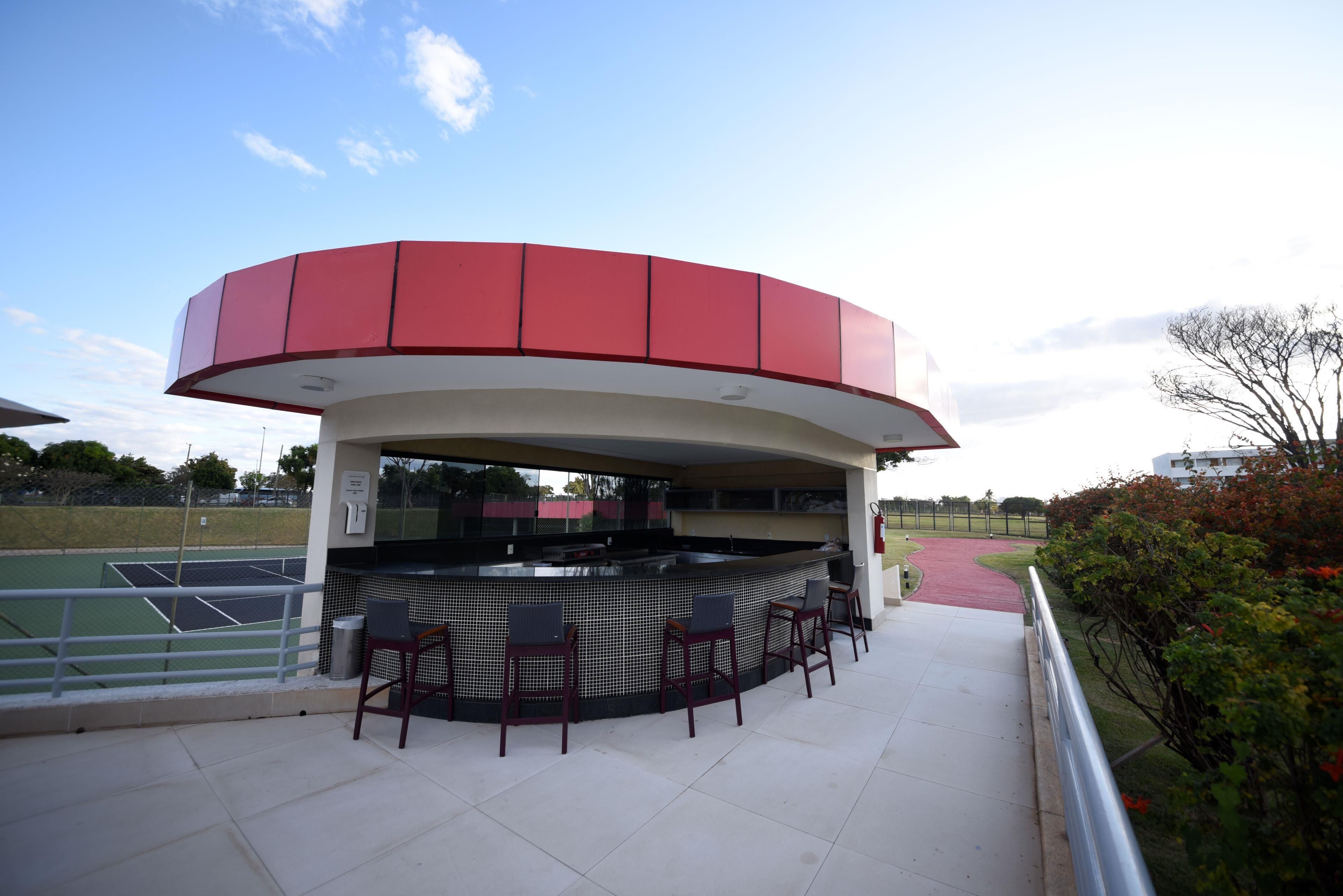 A covered outdoor bar area with a red and white structure, a tennis court in the background, and a paved walkway.