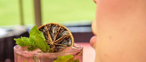 A person holding a tall glass with a red beverage, garnished with mint leaves and a dried orange slice.