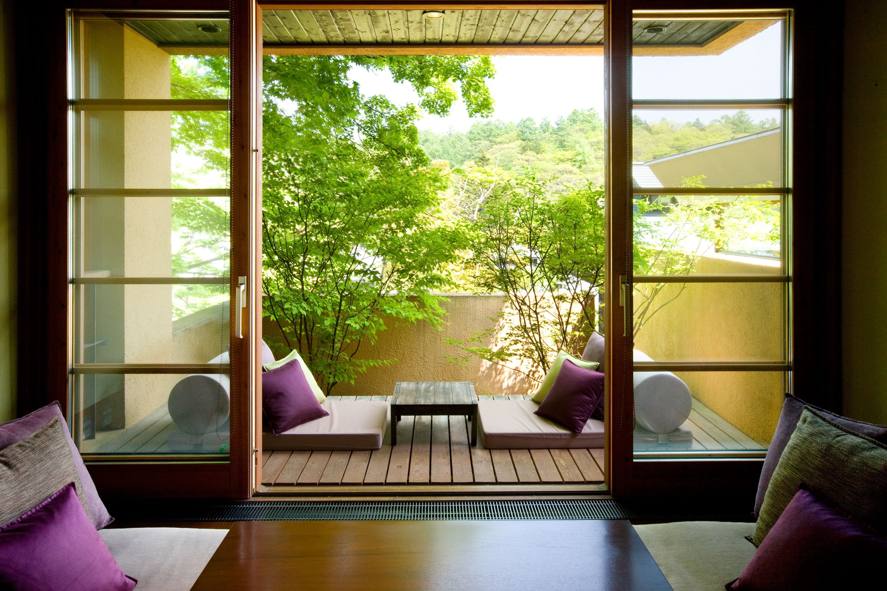 A room with a sliding glass door leading to a patio with a wooden table and two cushioned seating areas, each with a pillow.