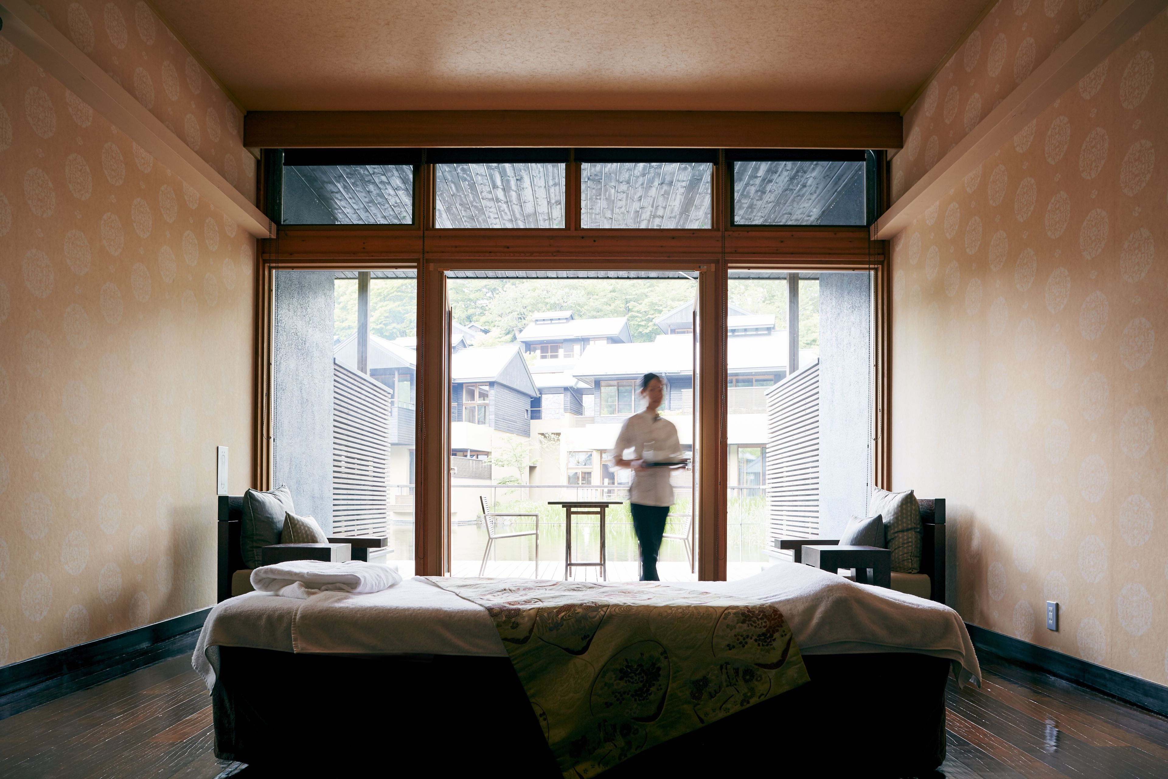A hotel room with a large bed, a small table, and a chair. There is a window with a view of buildings outside.