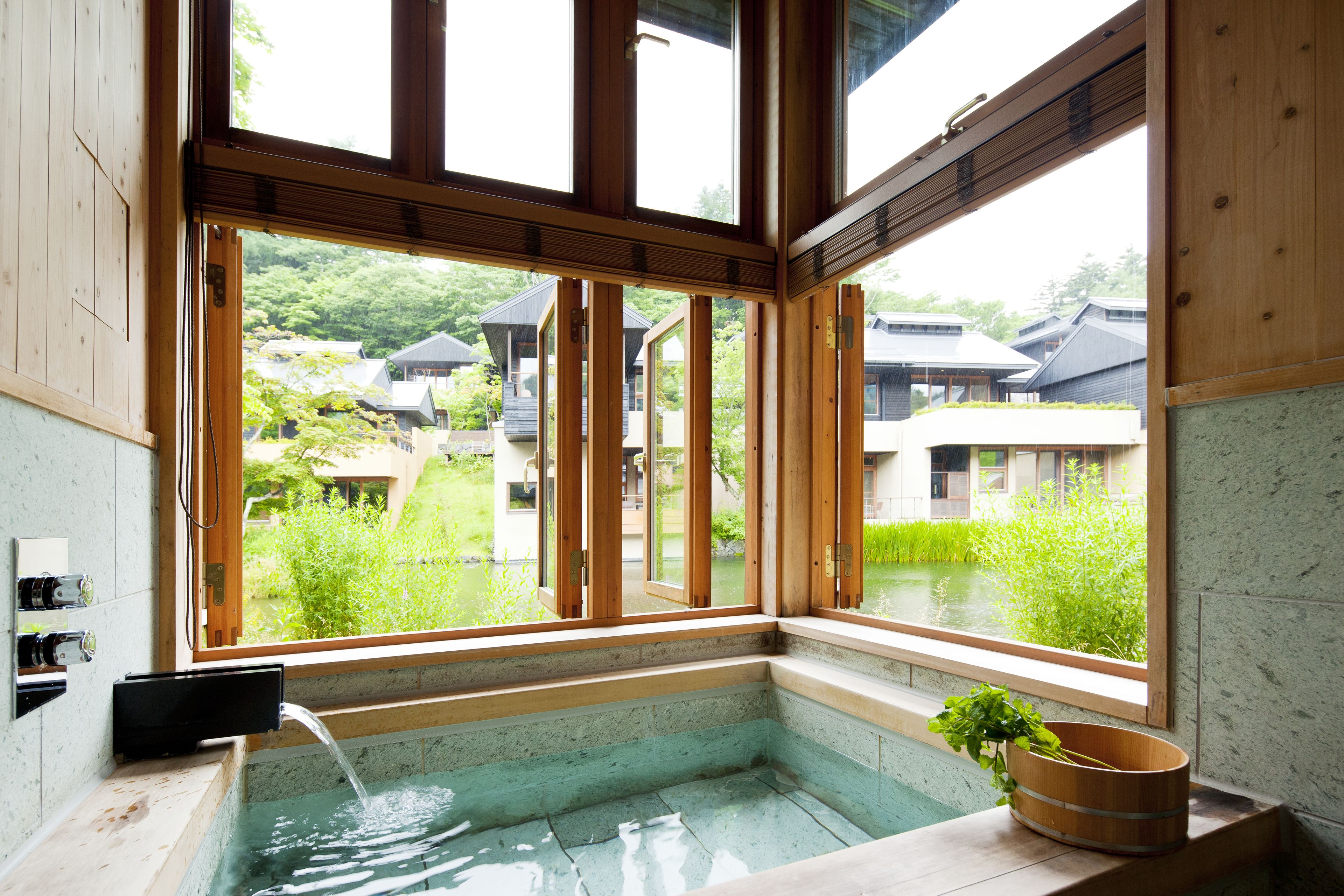 A traditional Japanese-style room with a wooden interior, a small onsen (hot spring) area, and a view of surrounding buildings and greenery.