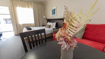 Une chambre d’hôtel avec un lit, un canapé rouge, une table à manger ornée d’un vase de fleurs et une fenêtre avec des rideaux.